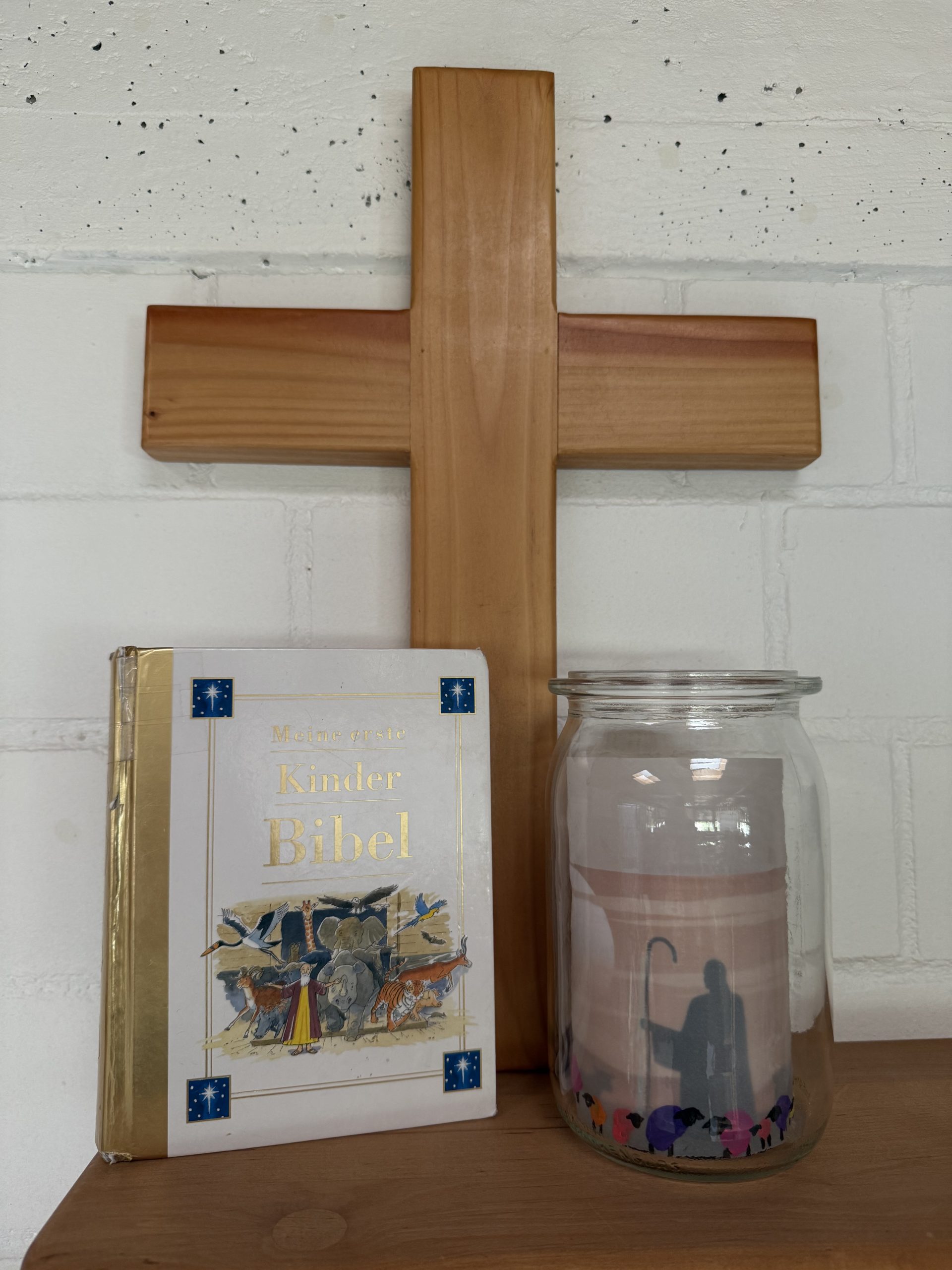Ein Bild auf dem eine Kinderbibel, ein Kreuz und ein großes Windlicht mit einem Schattenbild von Jesus als Hirte und bunte Fingerabdrücke der Kinder als Schafe gestaltet zu sehen ist.