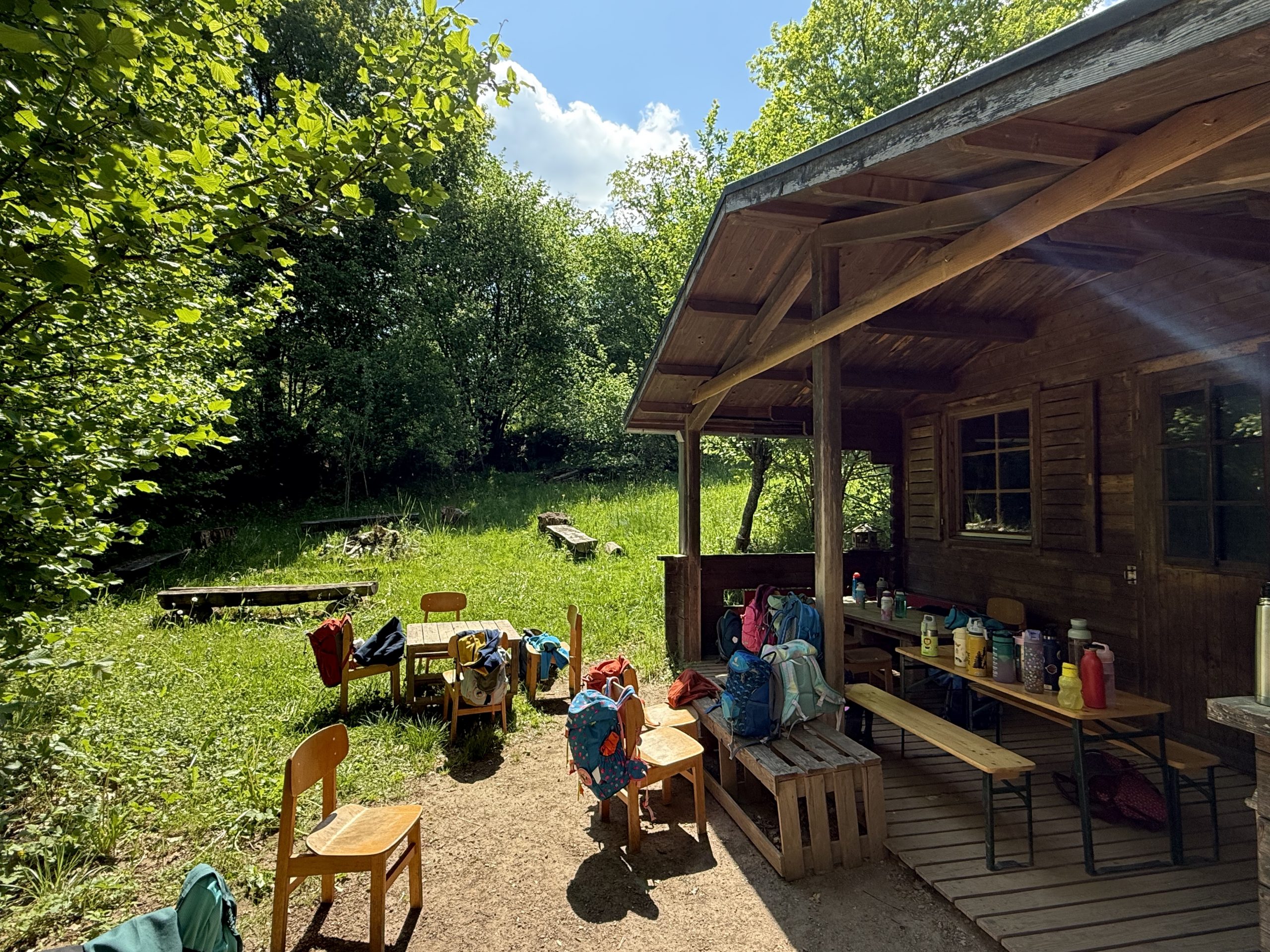 Sitzgelegenheit auf dem Schützenberg. Ein sonniger Ausschnitt unseres Platzes vor der Hütte, mit Stühlen, Tischen und den Rucksäcken der Kinder. Im Hintergrund unsere Baumstammbänke und das Gelände zum Spielen und Entdecken